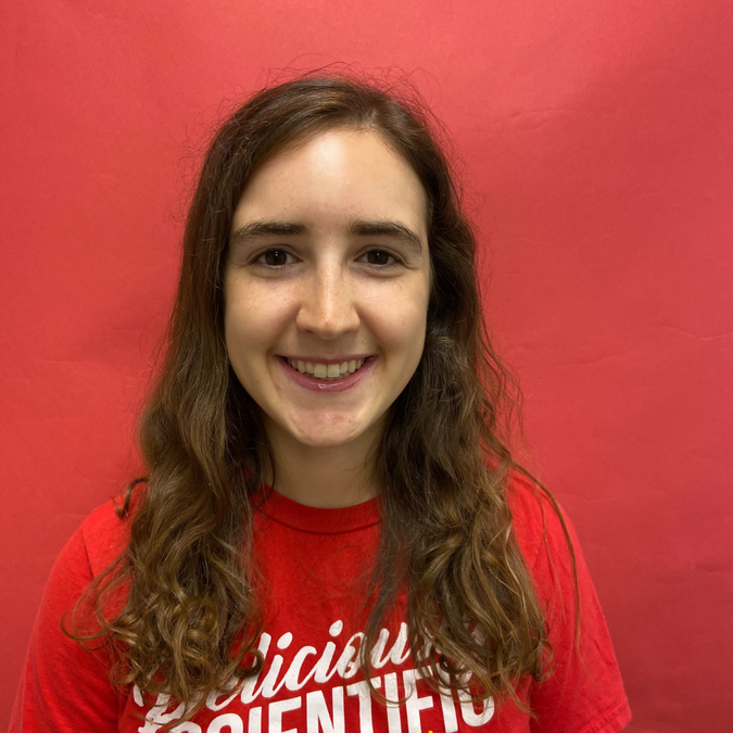 Brown haired female presenting person in a red shirt against a red background looking at the camera