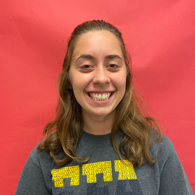 White presenting female with long brown hair looking at the camera wearing a grey shirt against a red background